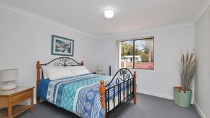 Een slaapkamer met een bed, nachtkastje, lamp, raam met uitzicht en een ingelijst schilderij aan de muur.