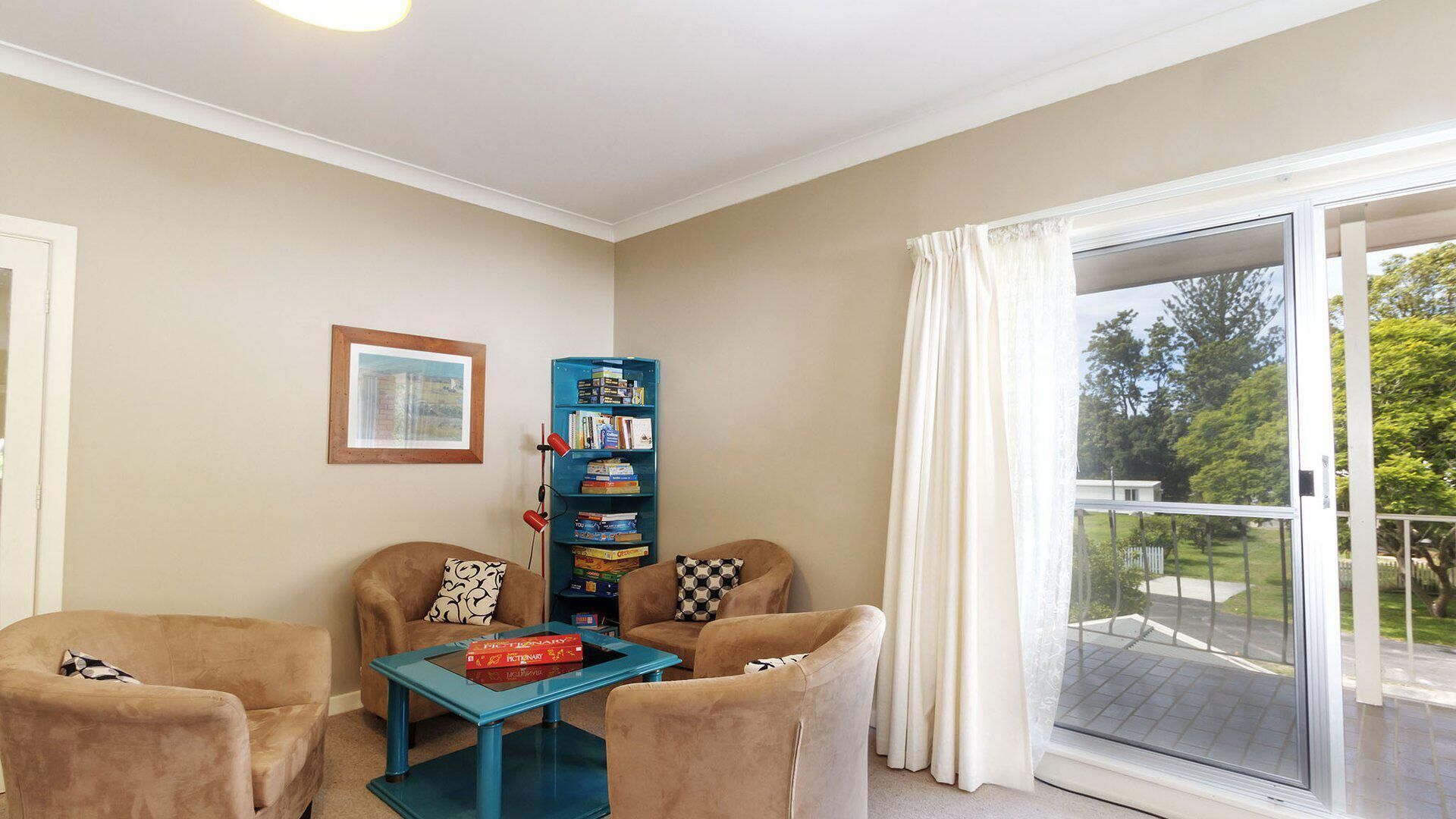 A living room with two armchairs, a coffee table, a bookshelf, a framed picture, and a sliding glass door to the outdoors.