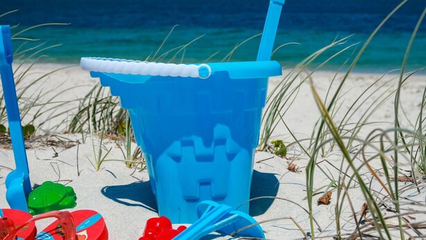 Un enfriador azul de playa con una pajita, sobre una playa arenosa con el mar al fondo.