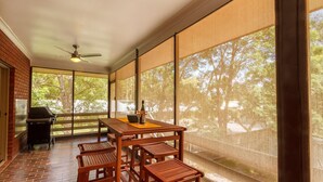 Un patio techado con mesa y sillas, un ventilador de techo y vistas a árboles a través de amplios ventanales.