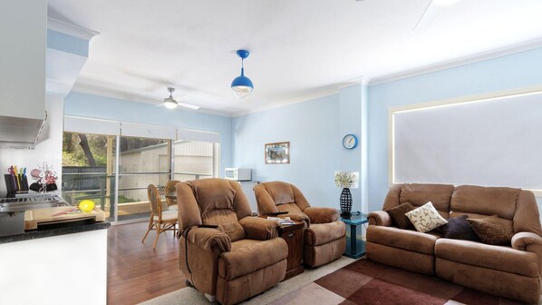 A living room with a sofa set, a dining area with a table and chairs, a ceiling fan, and a large window.