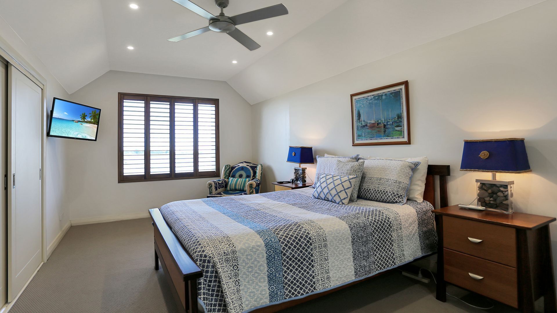 A bedroom with a bed, a ceiling fan, a window with blinds, a chair, a bedside table with a lamp, and a painting on the wall.