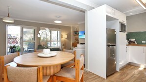 Eine moderne Küche mit Einbaukühlschrank, ein Essbereich mit rundem Holztisch und Stühlen sowie ein großes Fenster mit Jalousien.