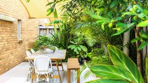 Eine Terrasse mit Holztisch und -stühlen, umgeben von üppiger Bepflanzung und einer Ziegelmauer.