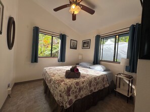 Ein Schlafzimmer mit einem Bett, einem Deckenventilator, zwei Fenstern mit Vorhängen, einem Nachttisch und einem an der Wand befestigten Regal.