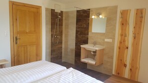 Une chambre avec une porte en bois, une douche avec un pare-douche en verre, un lavabo avec un miroir et un lit avec du linge de lit blanc.