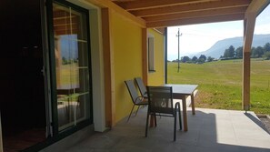 Une terrasse couverte avec une table et des chaises, une porte-fenêtre coulissante et une vue sur un champ verdoyant et des montagnes.