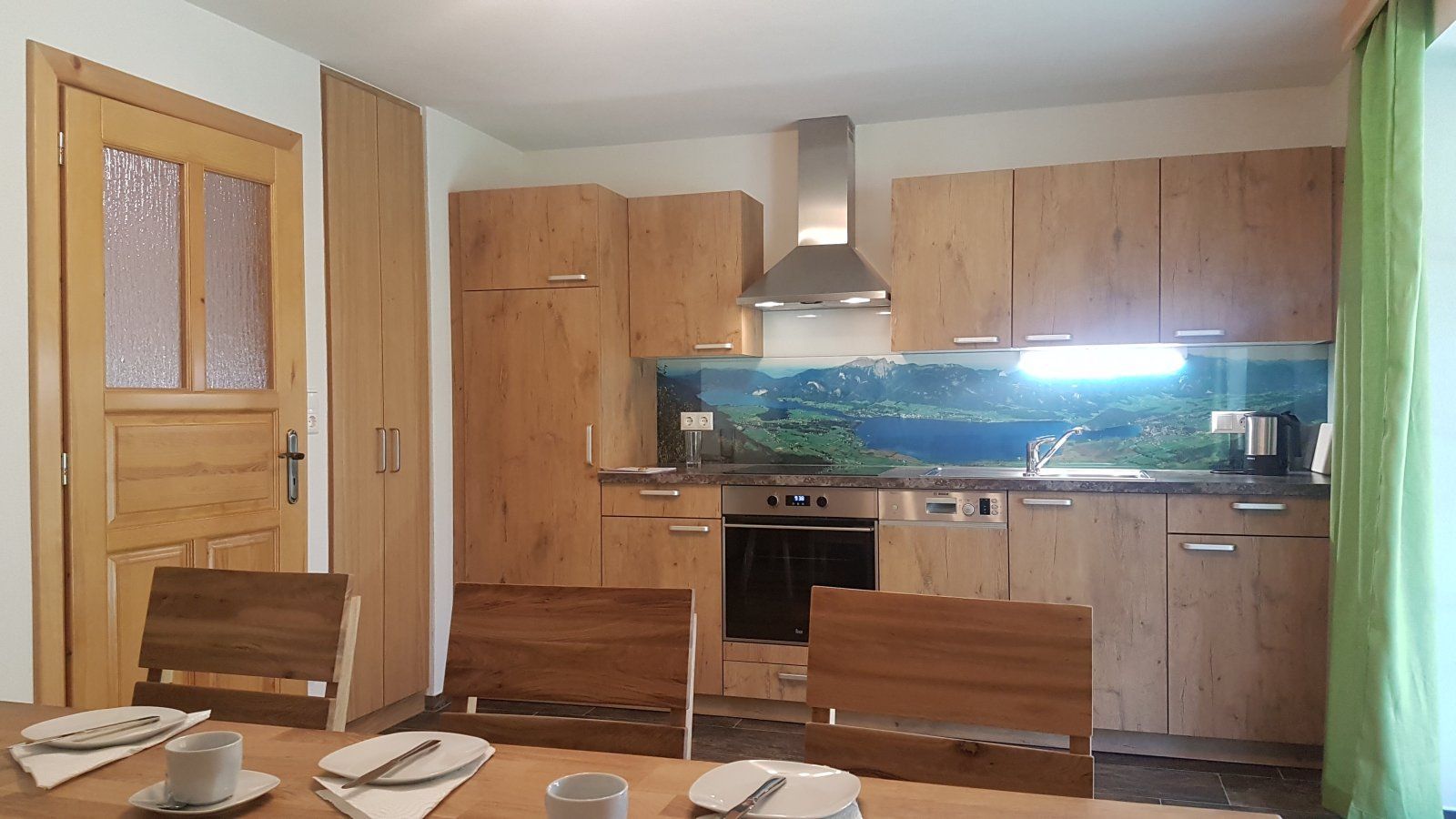Een keuken met houten kastjes, een fornuis en een eethoek met stoelen en een tafel gedekt voor twee personen.