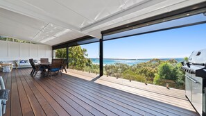 A covered outdoor deck with a wooden floor, a table with chairs, and a grill. - Belle Vue Beach House (Point Lookout)