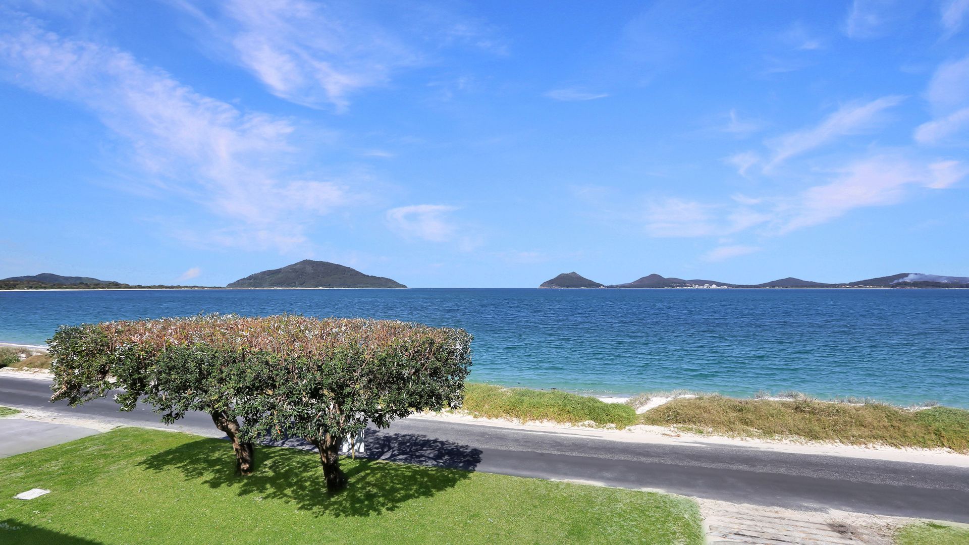 A coastal road with a grassy area and a tree, leading to a calm sea with distant islands.