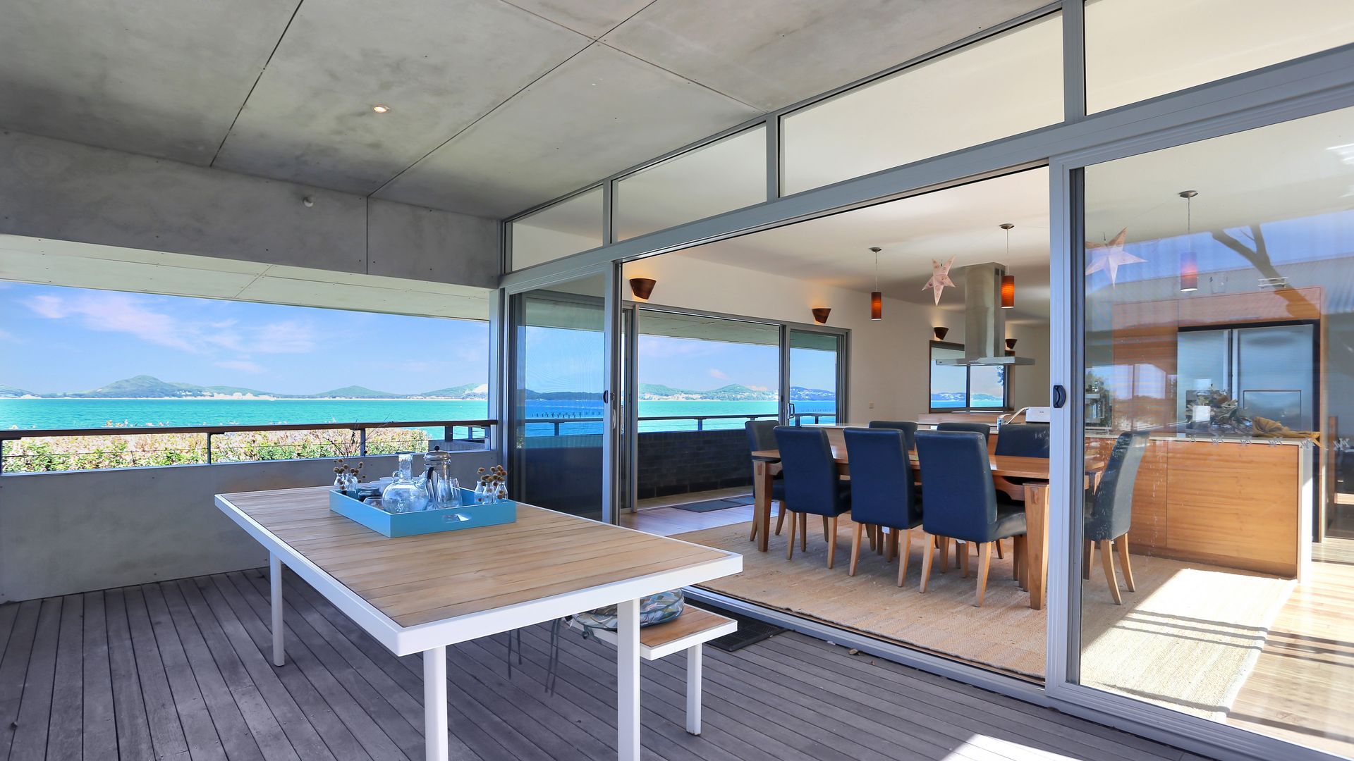 A modern outdoor dining area with a view of the sea, a wooden table with a tray of items, and a glass-enclosed dining space with chairs.