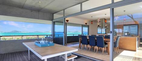 A modern outdoor dining area with a view of the sea, a wooden table with a tray of items, and a glass-enclosed dining space with chairs.