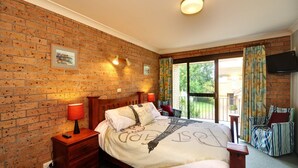 A bedroom with a brick wall, a bed with a Paris-themed pillow, a nightstand with a lamp, and a view of a garden through a sliding door. - Close To The Main Surf Beach (Hawks Nest)