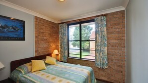 A bedroom with a brick wall, a bed with floral bedding, a window with curtains, and a painting on the wall. - Close To The Main Surf Beach (Hawks Nest)