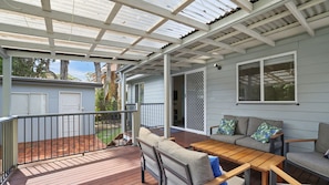 A covered outdoor deck with a wooden table and chairs, a sofa with cushions, and a sliding glass door leading to an interior space.