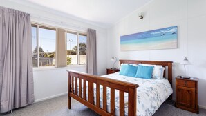 Ein Schlafzimmer mit einem hölzernen Bett, zwei Nachttischen, einem Beistelltisch, einem Fenster mit Vorhängen und einem Strandbild an der Wand.