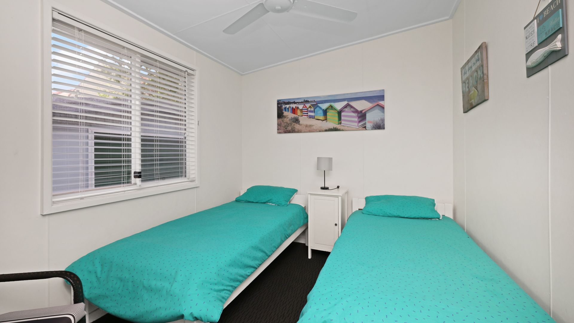 A room with two single beds, a small bedside table with a lamp, a window with blinds, and a wall with a painting.