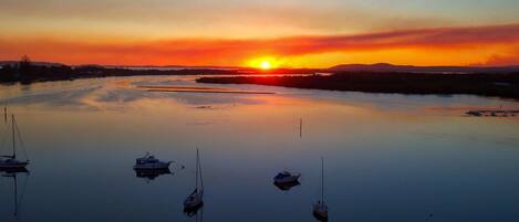 Una superficie acuática tranquila con varias embarcaciones ancladas, un atardecer en el fondo y una costa distante.