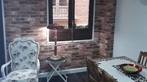 A room with a brick wall, a window, a table with chairs, a small table with a lamp, and a chair with floral upholstery. - IN THE HEART OF GRANADA - WIFI (Granada)