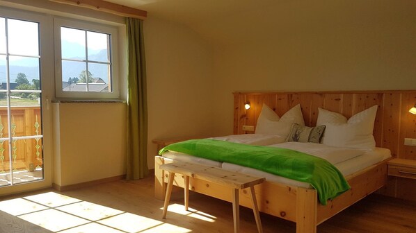A bedroom with a wooden bed, a bench, a window with curtains, and a view of the outdoors.