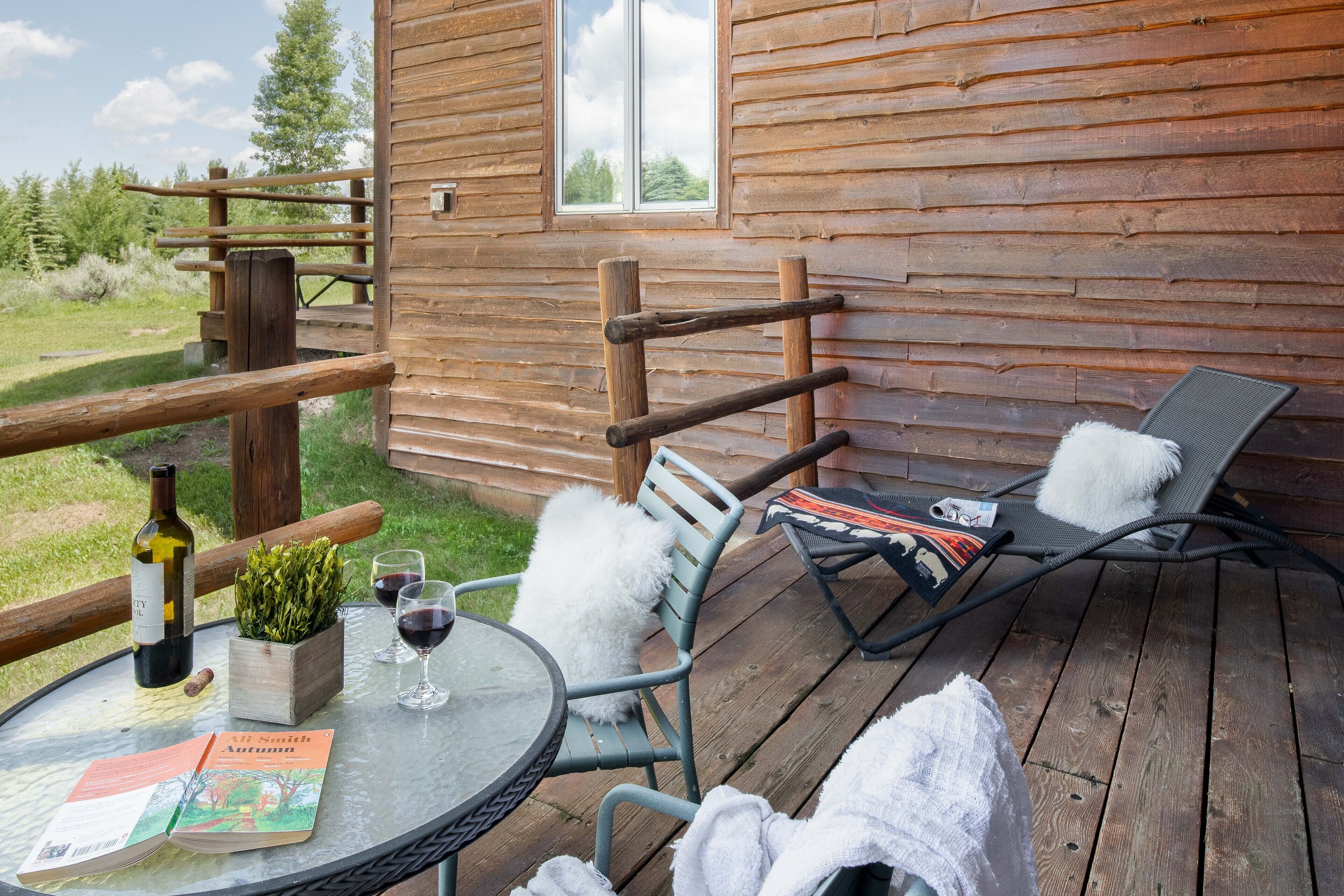 Una cabaña de madera con un balcón, una mesa con un libro, una botella de vino y una silla con una toalla.
