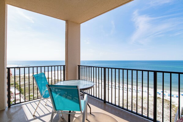 A balcony with a glass table and chairs overlooking a beach with a clear sky.