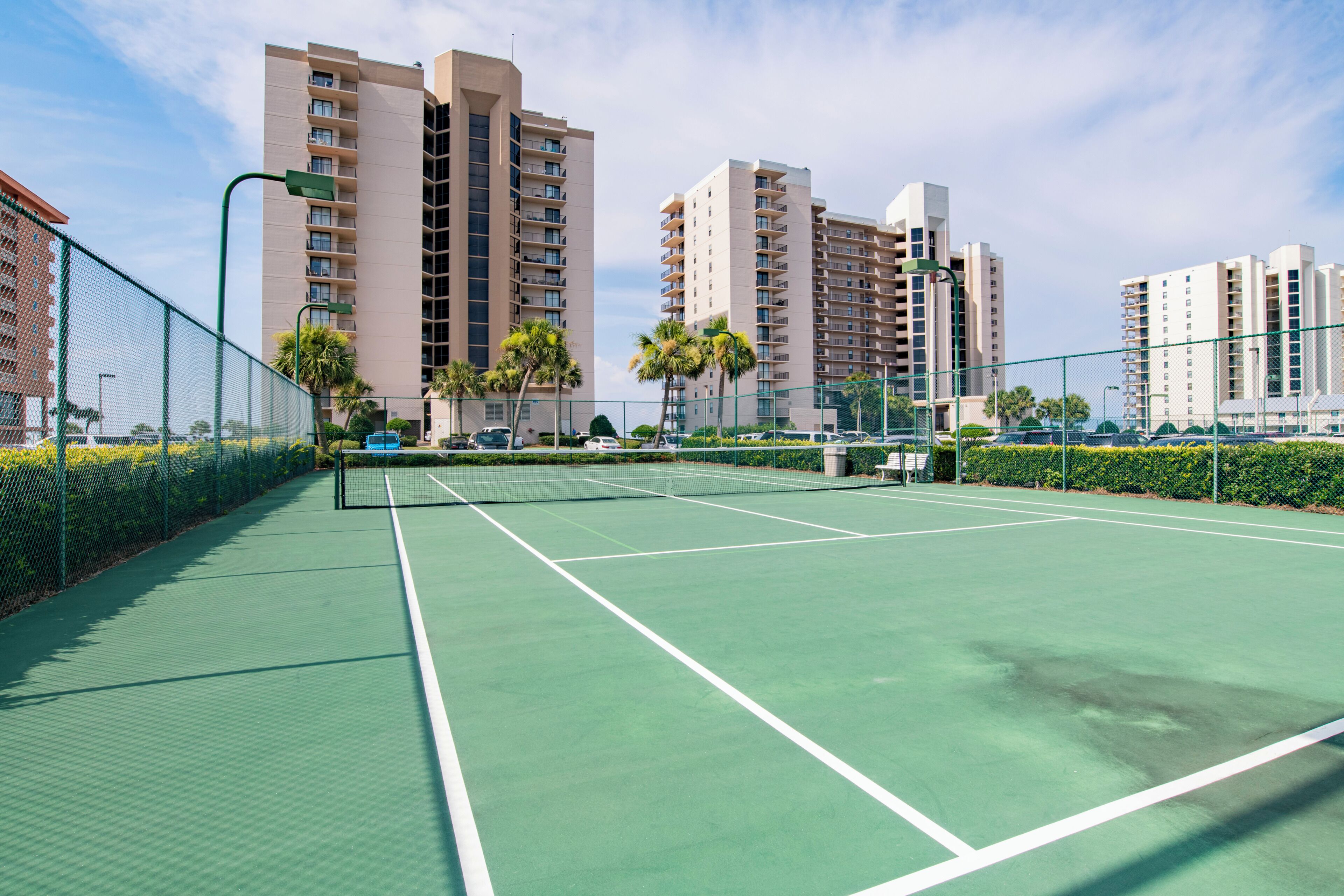 Un court de tennis avec un filet, entouré d’une clôture, et situé à côté d’immeubles d’habitation de grande hauteur.