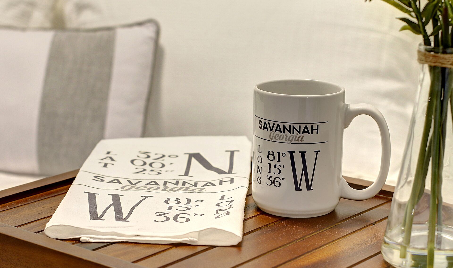 Une tasse blanche ornée de l’inscription « Savannah, Géorgie » et de coordonnées géographiques, une carte pliée sur laquelle figure le mot « Savannah », et un vase contenant des plantes vertes.