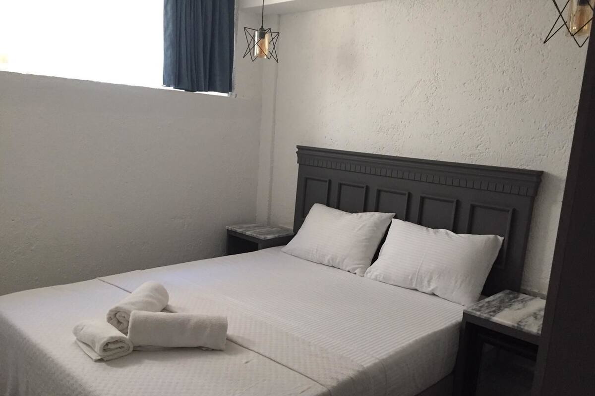 Una cama bien hecha con sábanas blancas y dos almohadas, una mesita de noche con una lámpara, y una ventana con cortinas azules.