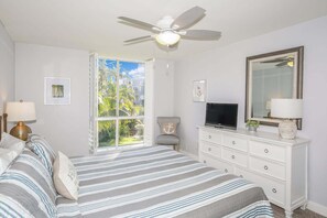 Een slaapkamer met een bed, een dressoir, een televisie, een plafondventilator en een raam met uitzicht op groen.