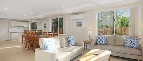 A spacious living area with a sofa, coffee table, and dining table. There is a kitchen in the background with white cabinets and a window.