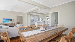 A spacious living area with a wooden dining table, comfortable seating, a television, and a kitchen in the background.