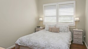 A bedroom with a bed, two bedside tables, a window with blinds, and a nightstand with a lamp.