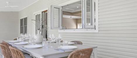 Un comedor con una larga mesa dispuesta para seis personas, paredes blancas y ventanas amplias con contraventanas abiertas.