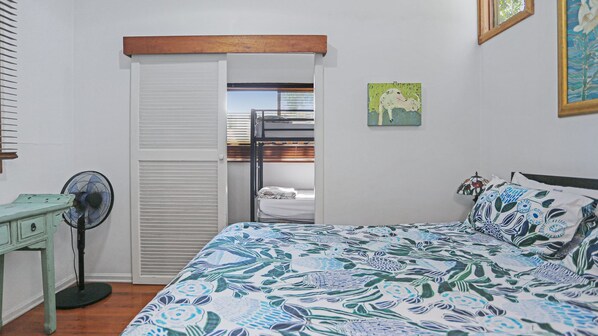 A bedroom with a bunk bed, a desk with a fan, a patterned bedspread, and a painting on the wall.