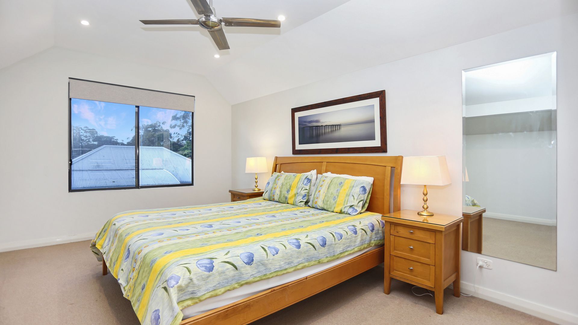 A bedroom with a bed, two bedside tables, a ceiling fan, a window with a view, and a framed picture on the wall.