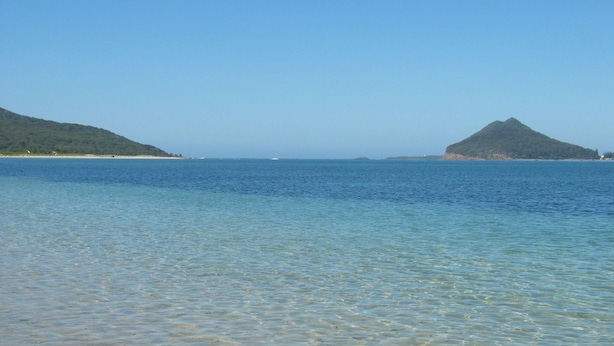 On peut clairement distinguer une zone côtière avec des eaux calmes, une plage de sable et deux collines verdoyantes en arrière-plan.