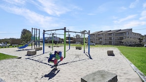 A playground with swings and a slide, surrounded by sand and grass, with a building in the background.