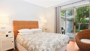Ein Schlafzimmer mit einem großen Bett, zwei Nachttischen mit Lampen, einem Korbstuhl und einer Glas-Schiebetür, die auf einen Balkon führt.