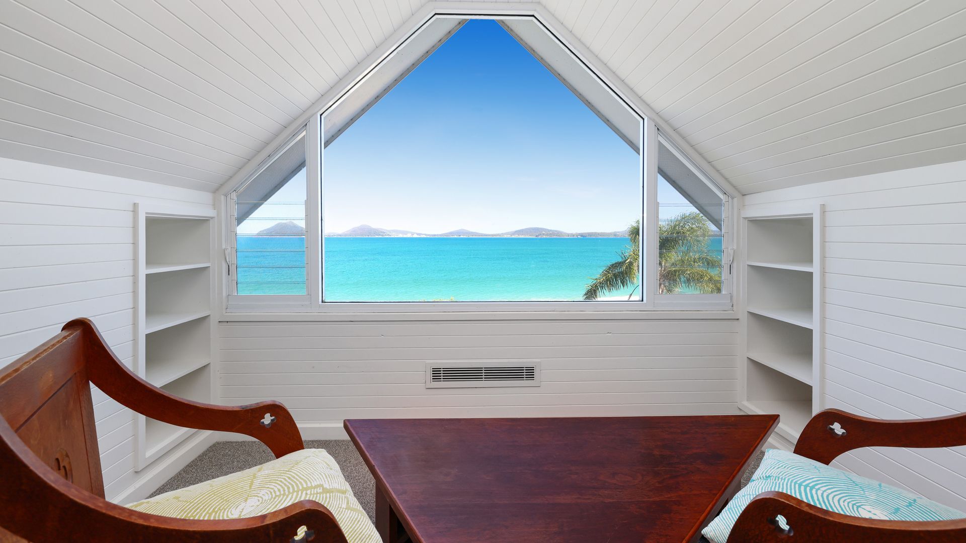 A room with a sloped ceiling, a large window with a view of the sea, and built-in wooden shelves.