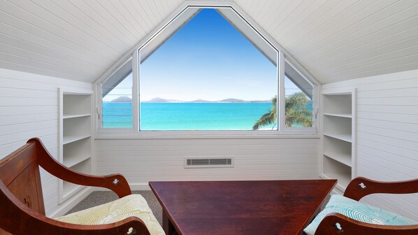 A room with a sloped ceiling, a large window with a view of the sea, and built-in wooden shelves.
