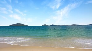 A beach with clear turquoise water, gentle waves, and distant islands under a clear sky.