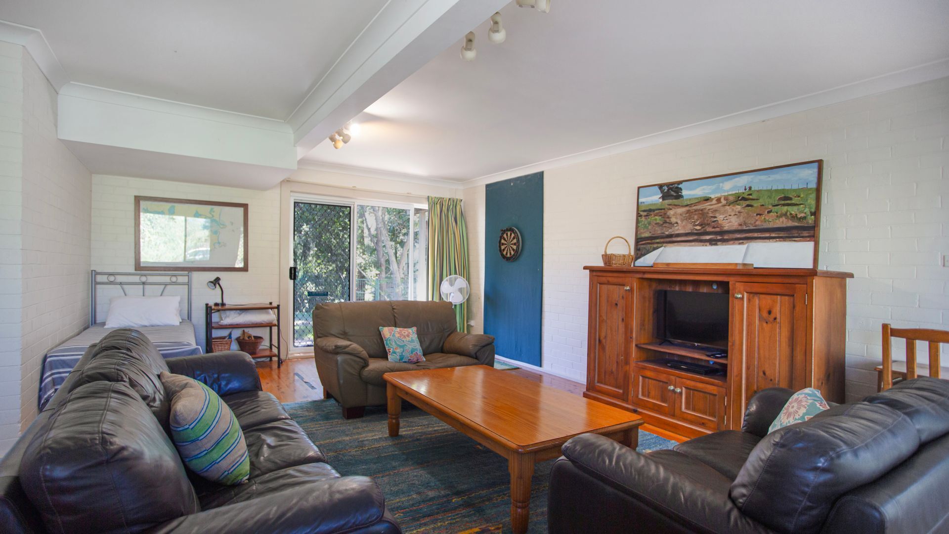 A living room with a sofa set, a wooden coffee table, a television on a wooden stand, a dartboard, and a painting on the wall.