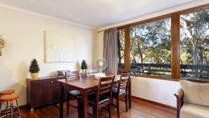 Un coin repas avec une table en bois, des chaises, un ventilateur et une vue sur les arbres à travers de grandes fenêtres.