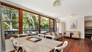 Una habitación amplia con una gran mesa de madera para comer, varias sillas y un sofá. Hay ventanas grandes que ofrecen vistas a árboles y un balcón.