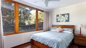 Une chambre avec une grande fenêtre, un lit recouvert d’une literie à motifs floraux, une table de chevet et un ventilateur de plafond.
