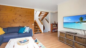 Un saló amb una paret de fusta, un sofà blau, una televisió que mostra una escena de platja i una escala.