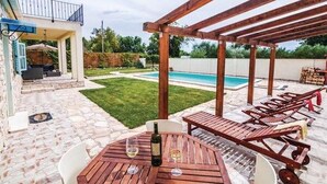 En overdækket udendørs terrasse med en pool, træloungestole og et bord med vinglas.