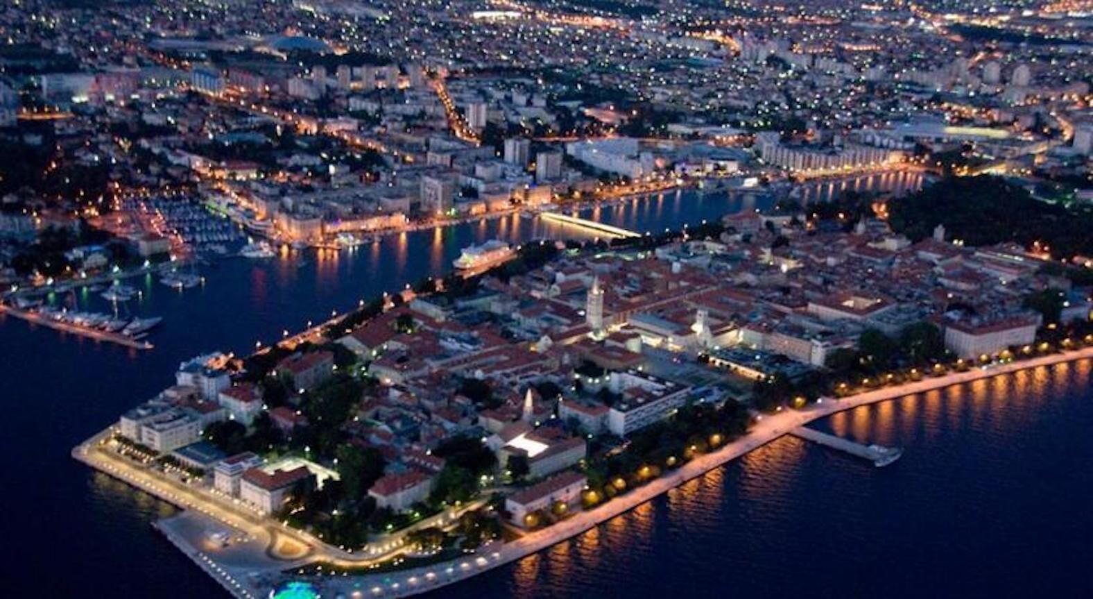An aerial view of a coastal city at night with illuminated streets and buildings, a marina with boats, and a prominent waterfront promenade.