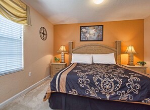A bedroom with a large bed, two bedside tables with lamps, a painting on the wall, and a window with blinds.